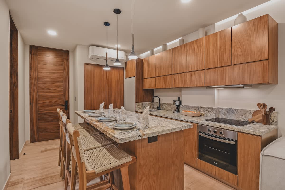 Open-concept kitchen and dining area featuring a long granite island with bar seating, light wood cabinetry, and integrated appliances, with an air conditioning unit visible above a wooden door.
