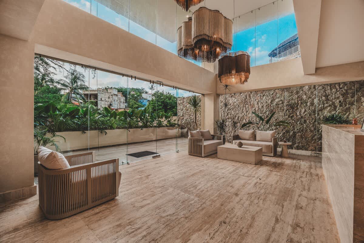 A grand, high-ceilinged lobby with large glass walls offering views of lush greenery, featuring modern seating areas with light-colored sofas and armchairs, and striking multi-tiered chandeliers.