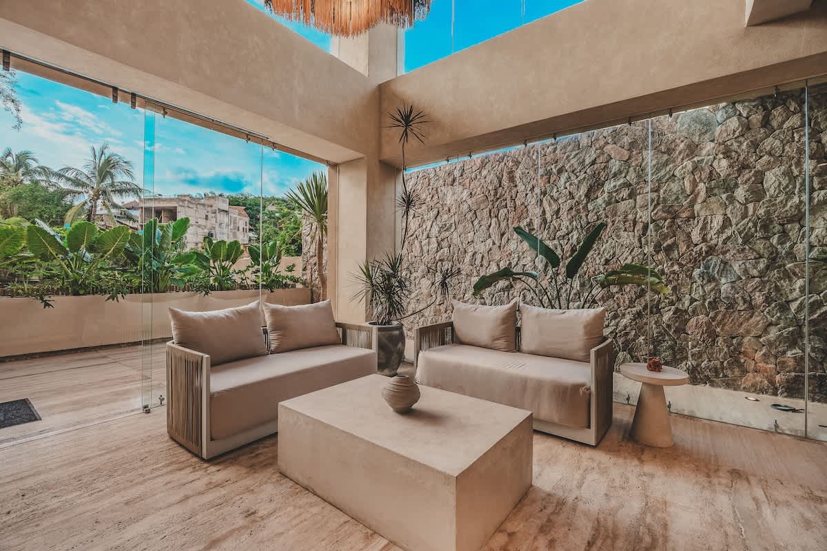 A close-up of the modern lobby seating area, showing two light-colored sofas and a rectangular coffee table, set against a textured stone accent wall and large glass windows overlooking tropical plants.