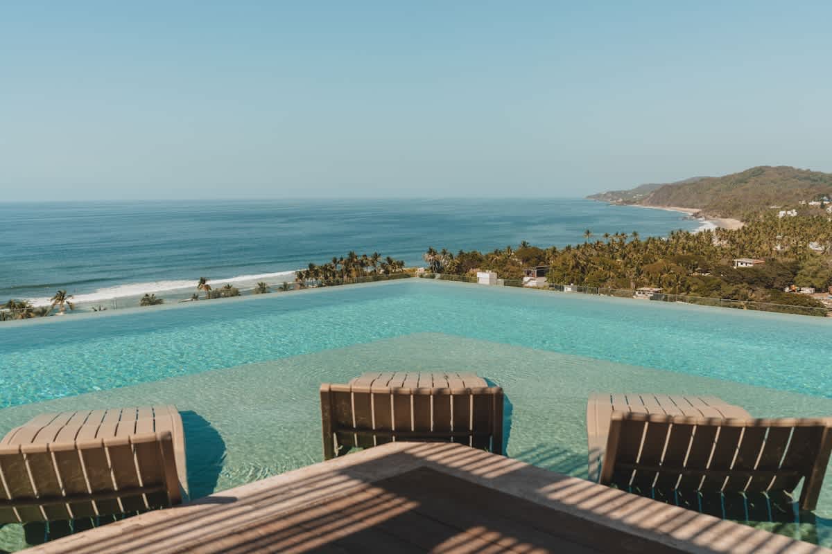 An expansive infinity pool with clear blue water features three submerged lounge chairs, offering breathtaking panoramic views of the ocean and a lush coastline under a clear sky. The pool edge blends seamlessly with the horizon.