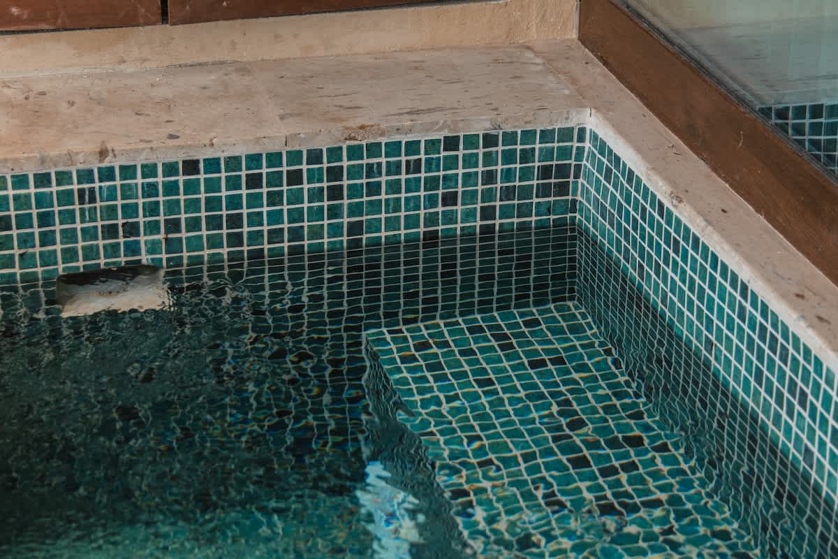 A close-up view of a square hot tub with dark teal mosaic tiles, showing submerged steps and bubbling water, set within a light-colored stone surround.