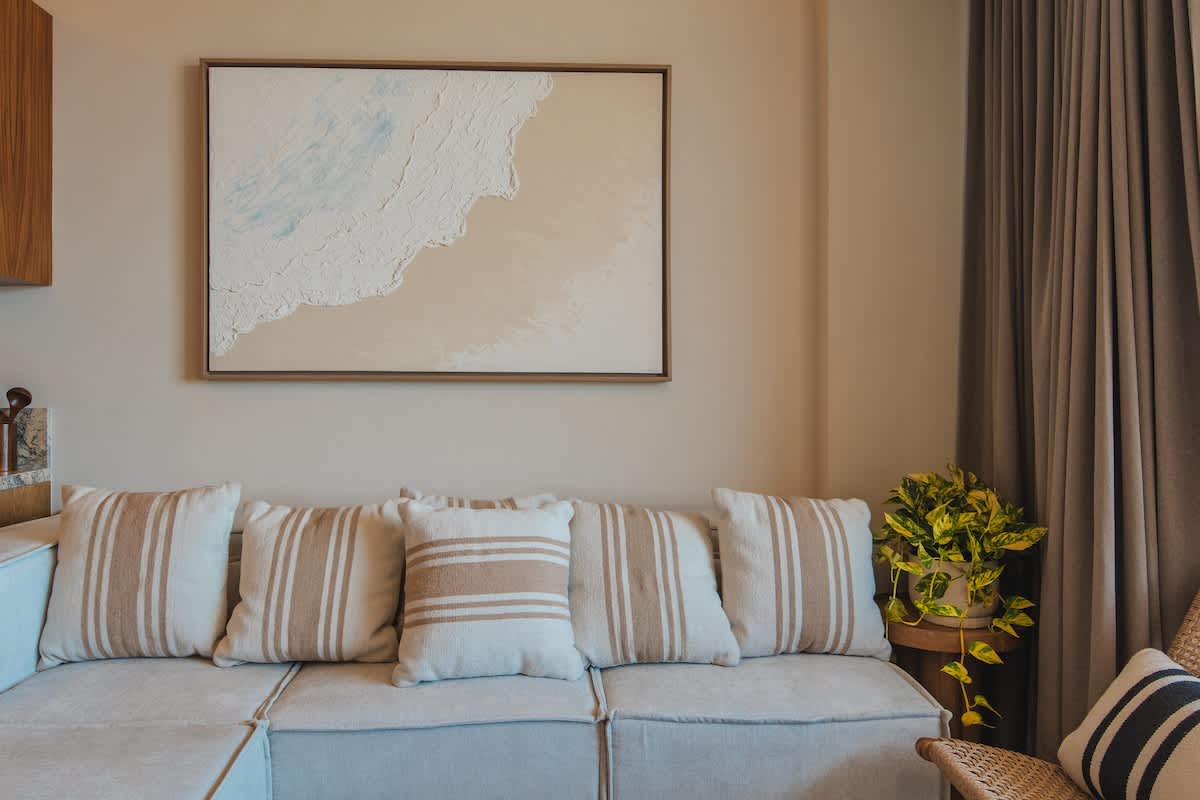 A cozy living room with a light-colored sofa adorned with striped throw pillows, a framed abstract artwork depicting a beach scene, and a vibrant green plant in the corner.
