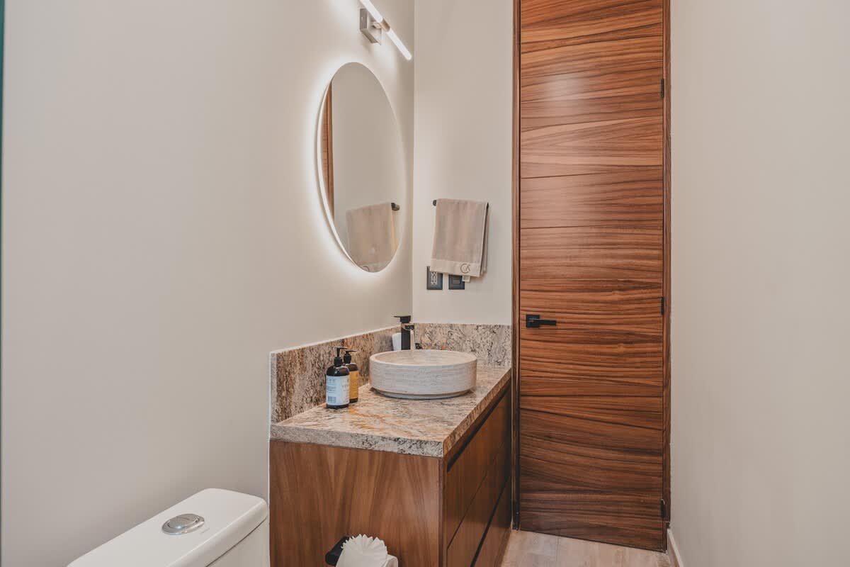 A modern bathroom featuring a light-colored stone vessel sink on a granite vanity, a backlit round mirror, and a white toilet partially visible on the left, with a tall wooden door on the right.