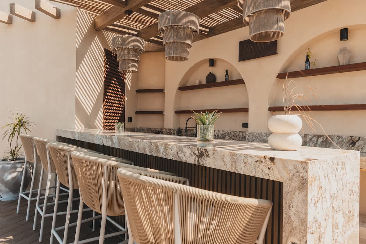 Stylish outdoor bar area with a long, veined marble countertop and several woven bar stools, set under a slatted wooden pergola with decorative pendant lights.