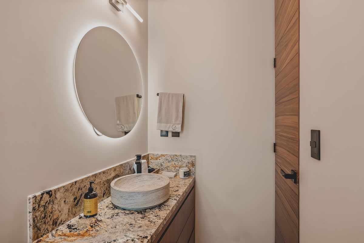 A close-up of the bathroom vanity highlights a textured vessel sink on a speckled granite countertop, illuminated by a round backlit mirror. Soap and rolled towels are neatly arranged.