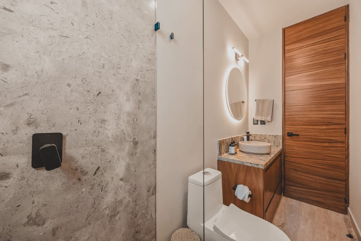 A stylish bathroom with a stone-tiled shower enclosure on the left, a white toilet, and a vanity featuring a vessel sink, granite countertop, and a backlit round mirror, all next to a tall wooden door.