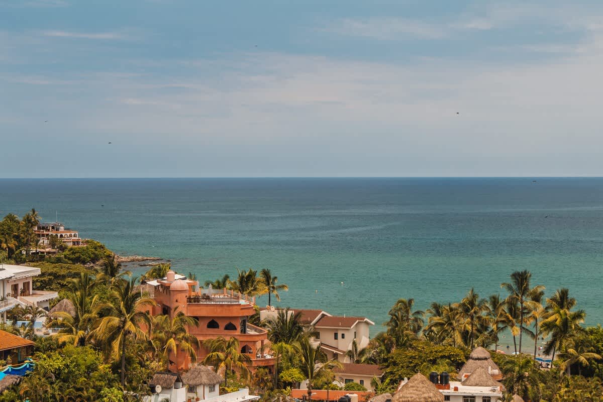 A scenic coastal view showcasing the vast blue ocean meeting a shoreline dotted with colorful houses and lush palm trees, under a bright sky with scattered clouds.
