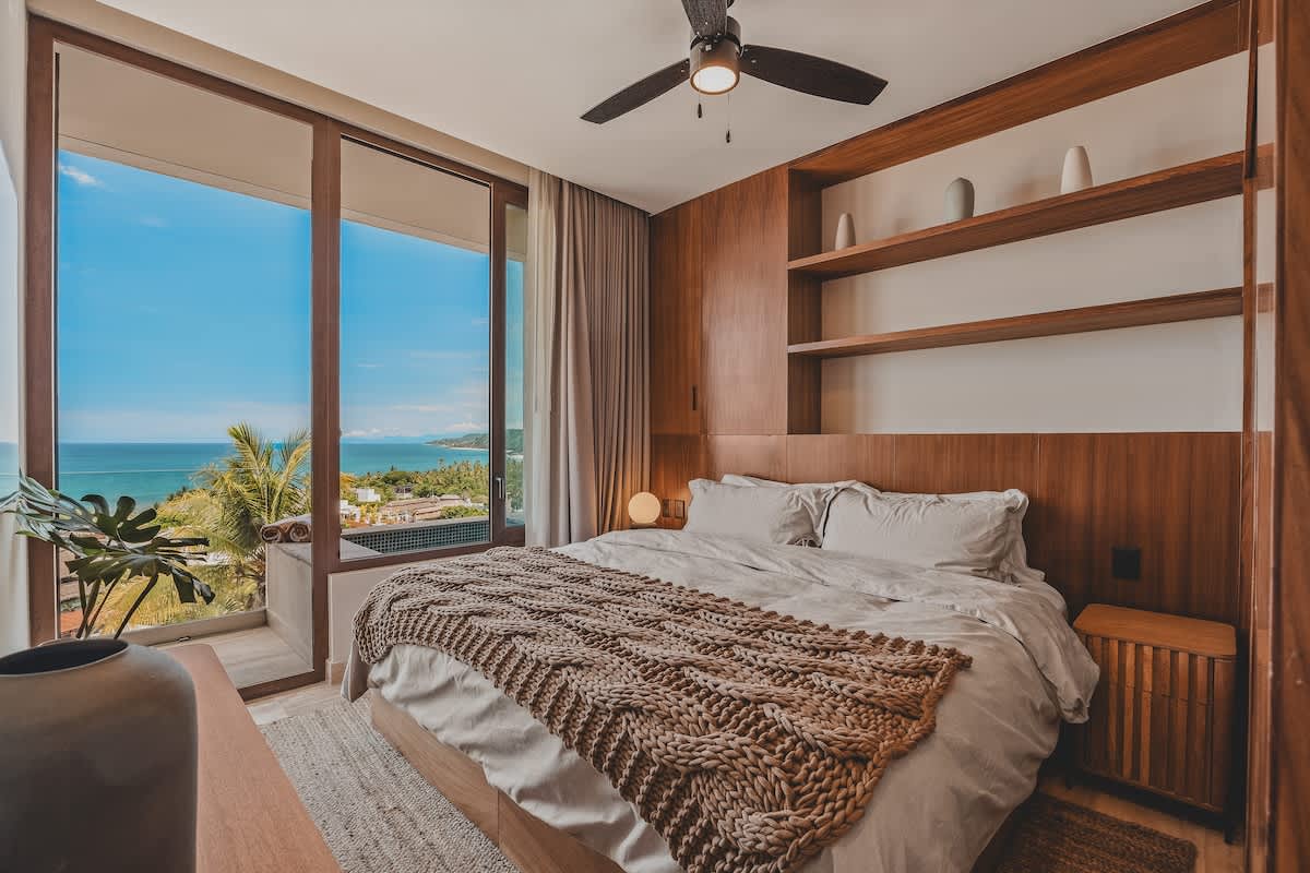 This bedroom features a large bed with a textured throw, built-in wooden shelving, and a ceiling fan. Sliding glass doors open to a balcony with a private hot tub and a breathtaking ocean vista.