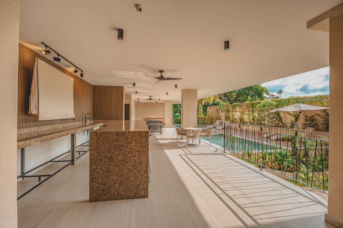 An open-air common area features a modern kitchen island with a granite-like countertop, a pool table, and a seating area, all overlooking a lush outdoor space with a swimming pool. The ceiling has recessed lighting and a fan.