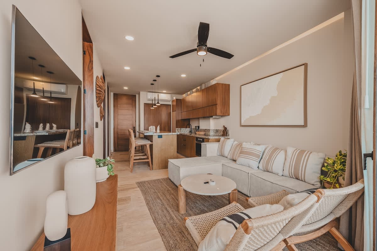 Bright living room with a cream sectional sofa, two woven lounge chairs, a round coffee table, and a large flat-screen TV, offering a glimpse of the kitchen and a stunning ocean view.