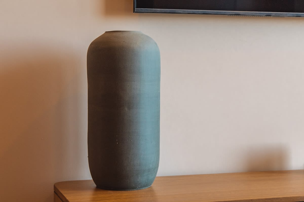 A close-up of a tall, cylindrical blue-grey ceramic vase resting on a wooden console table against a light-colored wall, showcasing its smooth texture.