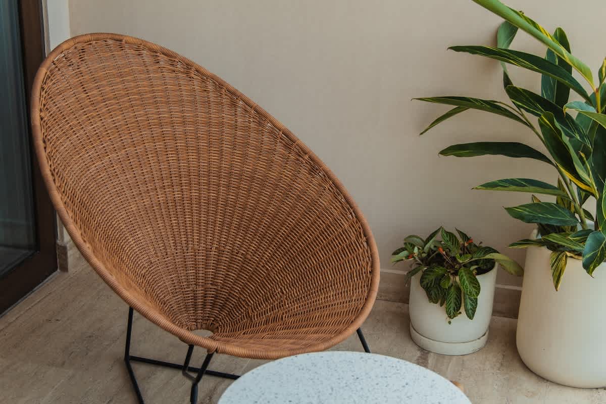 A modern, oval-shaped wicker chair with a black metal frame sits on a light-colored tiled balcony next to two potted plants against a neutral wall.