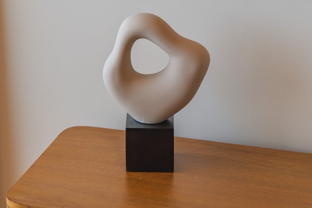A modern, abstract white sculpture with a smooth, organic, and fluid shape, resting on a dark square base atop a light wooden surface.