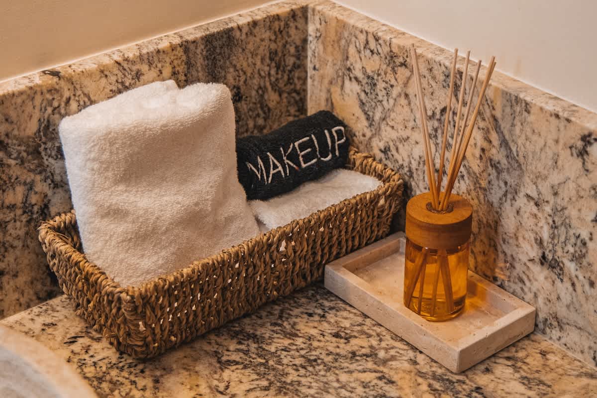 A close-up of a bathroom counter featuring a woven basket with white and black "MAKEUP" towels, a reed diffuser with amber liquid on a light stone tray, all set against a speckled granite countertop and backsplash.