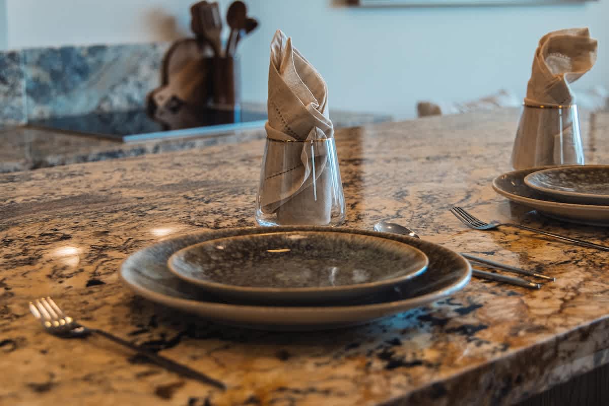 A dining area with a granite countertop serving as a table, set with two place settings featuring dark grey and beige ceramic plates, silver forks, and spoons. A clear glass with a folded beige napkin stands upright, adding a touch of elegance.