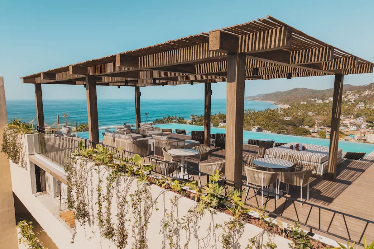 Wide shot of the luxurious rooftop deck with various seating arrangements under a wooden pergola, bordering an infinity pool, all set against a stunning backdrop of the ocean and a verdant coastal town.