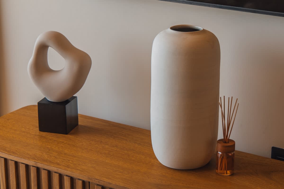 A wooden console table holds two decorative items: a beige abstract sculpture on a black base and a tall, matte white ceramic vase. A reed diffuser with amber liquid adds a subtle scent to the modern living space.