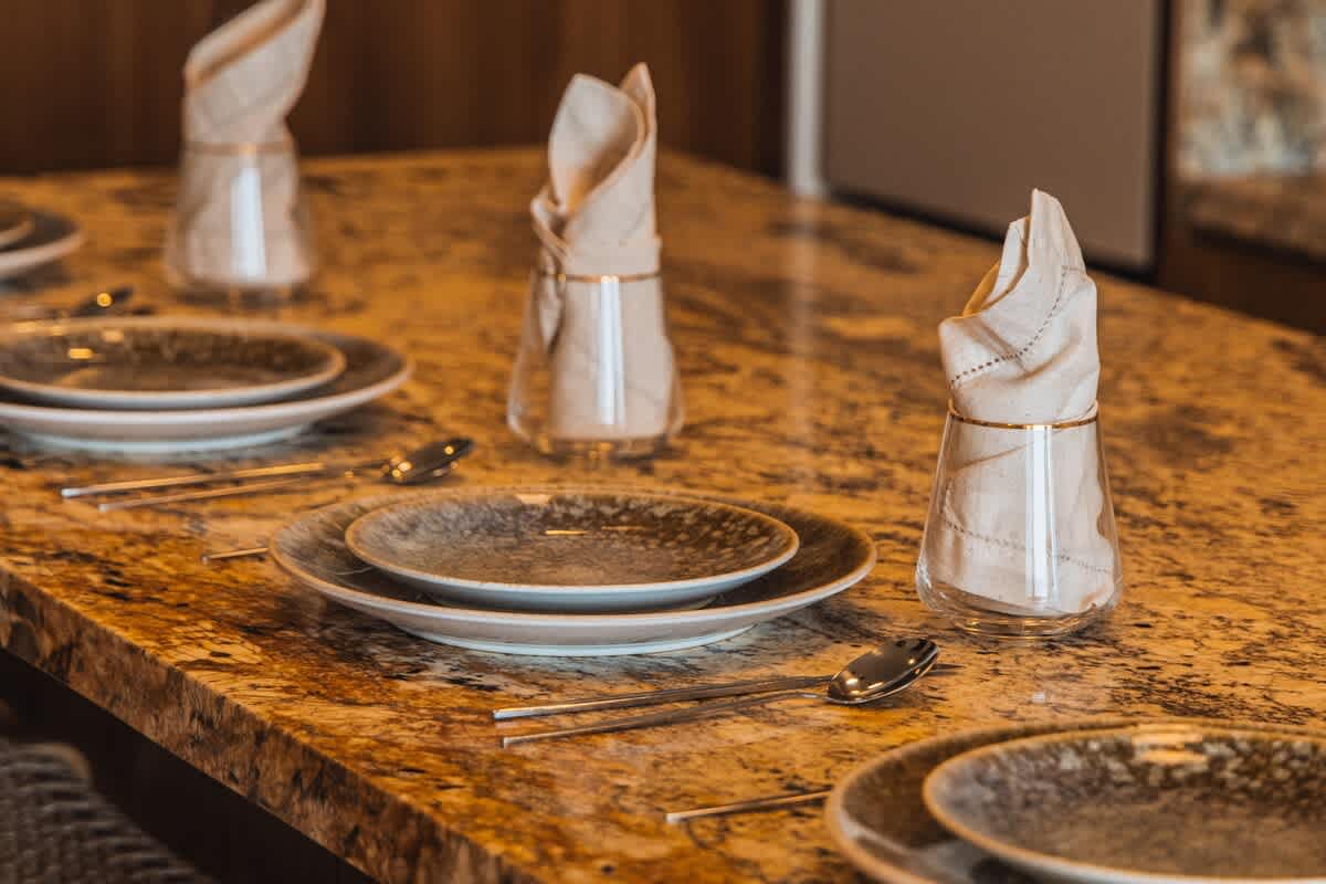 A wider view of the dining area, showcasing multiple place settings on a polished granite countertop. Each setting includes dark ceramic plates, silverware, and a glass with a neatly folded beige napkin, ready for a meal.