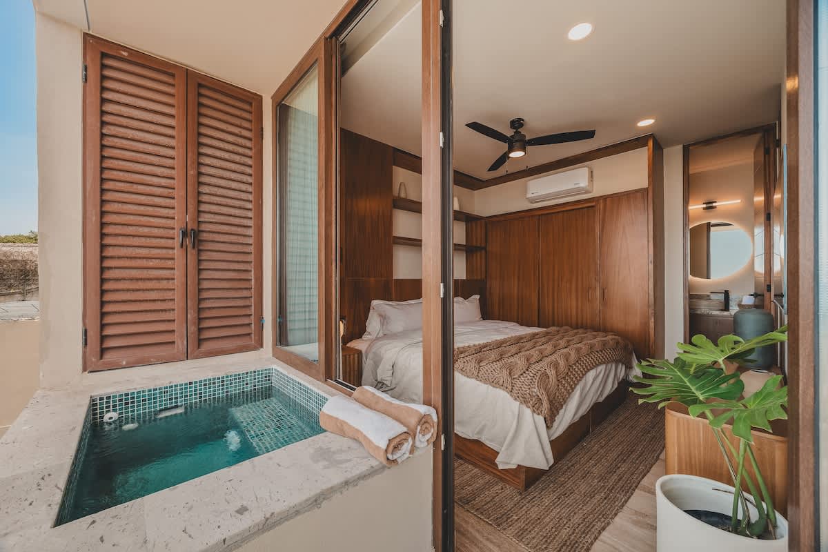 A bedroom with a large bed and wooden built-ins offers direct access to a balcony featuring a private hot tub. The view extends to the ocean, framed by open sliding doors and closed wooden shutters.