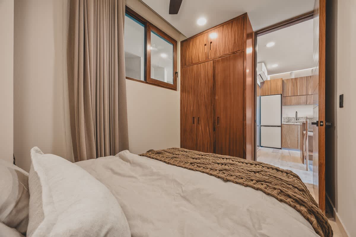 View from the bedroom showing a wooden built-in wardrobe and a glimpse into an adjacent modern kitchen with light wood cabinetry and a refrigerator.