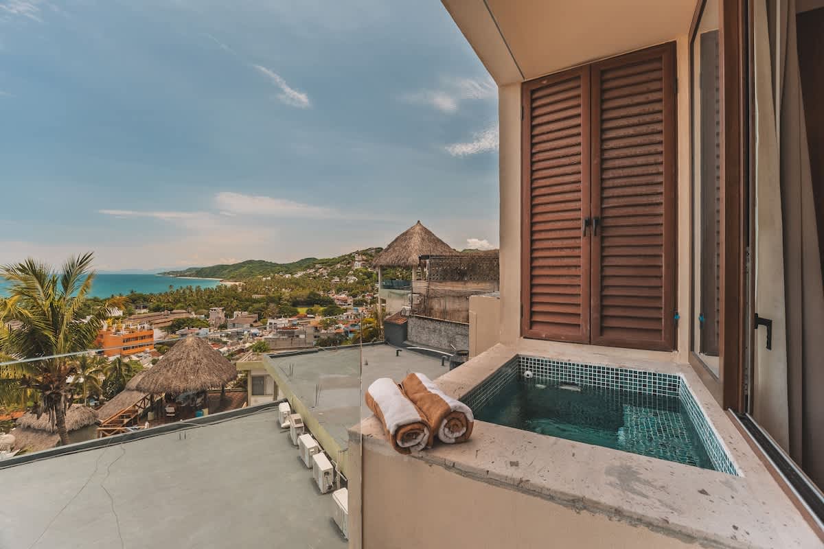 Outdoor private hot tub with blue mosaic tiles on a balcony, offering a scenic view of a coastal town and distant mountains, with rolled towels placed nearby.