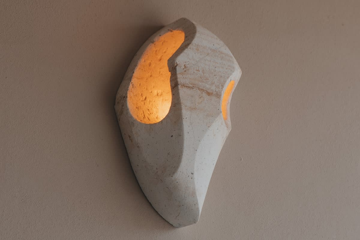 A unique, sculptural wall sconce made of light-colored, textured material emits a warm, soft glow from its organic-shaped openings against a neutral wall.
