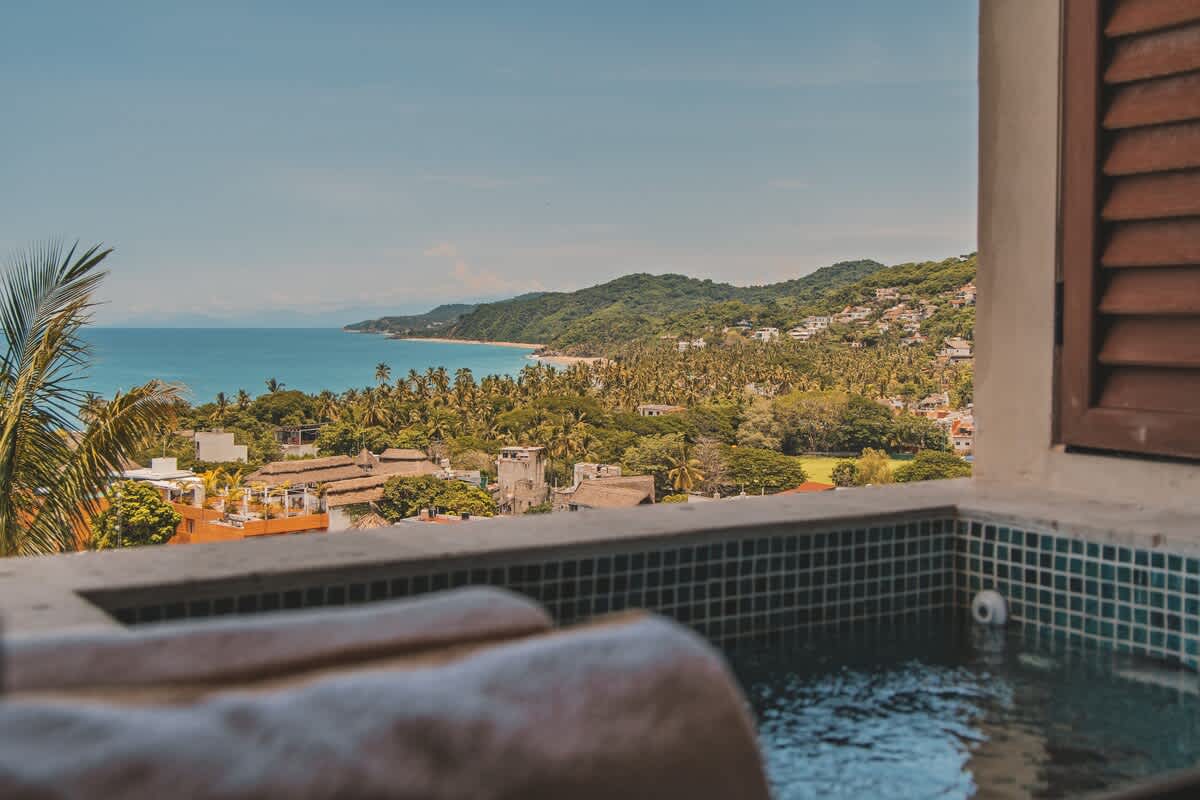 A hot tub with teal mosaic tiles offers panoramic views of a coastal town, lush green hills, and the expansive blue ocean under a clear sky.