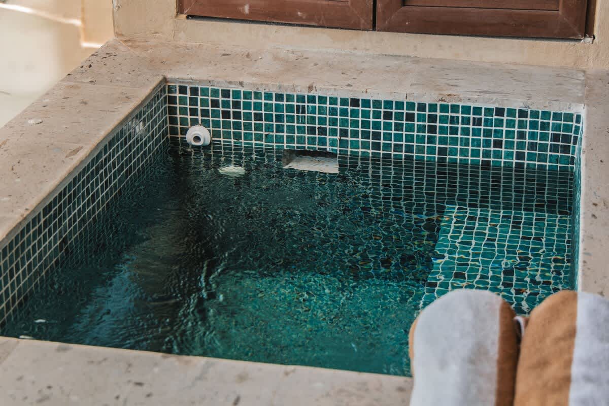 A square hot tub with dark teal mosaic tiles is filled with bubbling water, set into a light-colored stone surround, with two folded towels visible in the foreground.