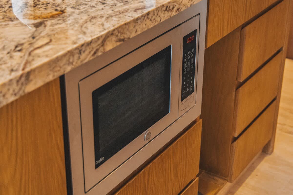 A built-in stainless steel microwave oven is seamlessly integrated into light wood cabinetry beneath a granite countertop. The microwave features a digital display and various control buttons, blending functionality with modern kitchen design.