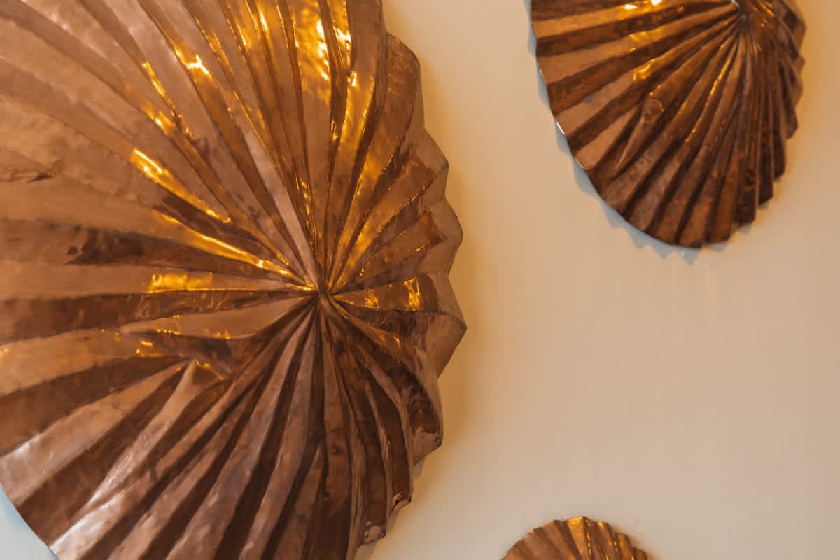 A close-up of a large, textured copper wall art piece, shaped like a fanned shell or leaf, with intricate radiating folds reflecting light.