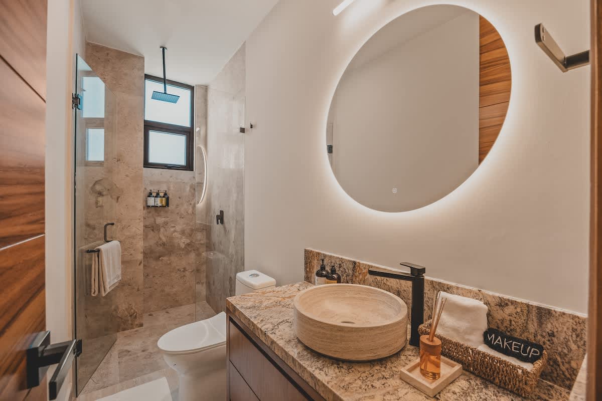 Modern bathroom featuring a circular backlit mirror above a vessel sink set on a granite vanity, with a spacious walk-in shower and light-colored tiled walls.