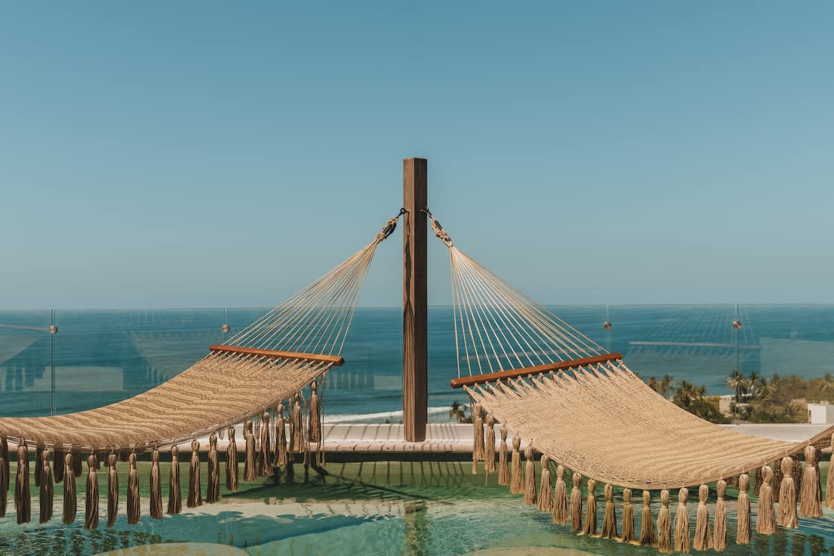Two woven hammocks with tassels are suspended over a clear blue infinity pool, offering a serene ocean view under a bright sky. The hammocks are attached to a central wooden post and have wooden spreader bars.