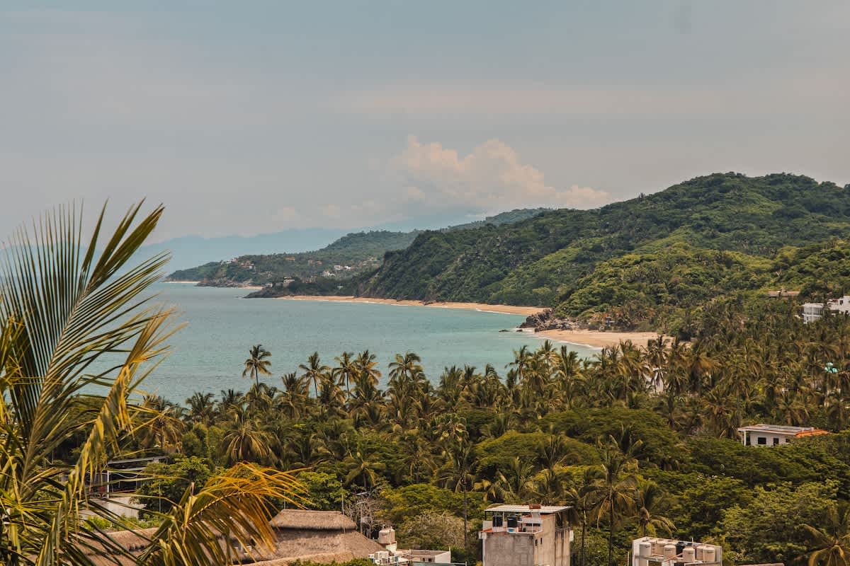 A panoramic view of a tropical coastline with a calm turquoise ocean, sandy beaches, and dense green hills covered in palm trees, with scattered buildings visible below.