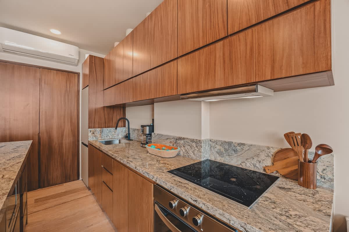 Modern kitchen counter with light granite, an integrated black induction cooktop, and a stainless steel oven, complemented by warm wood cabinetry and a sleek range hood.