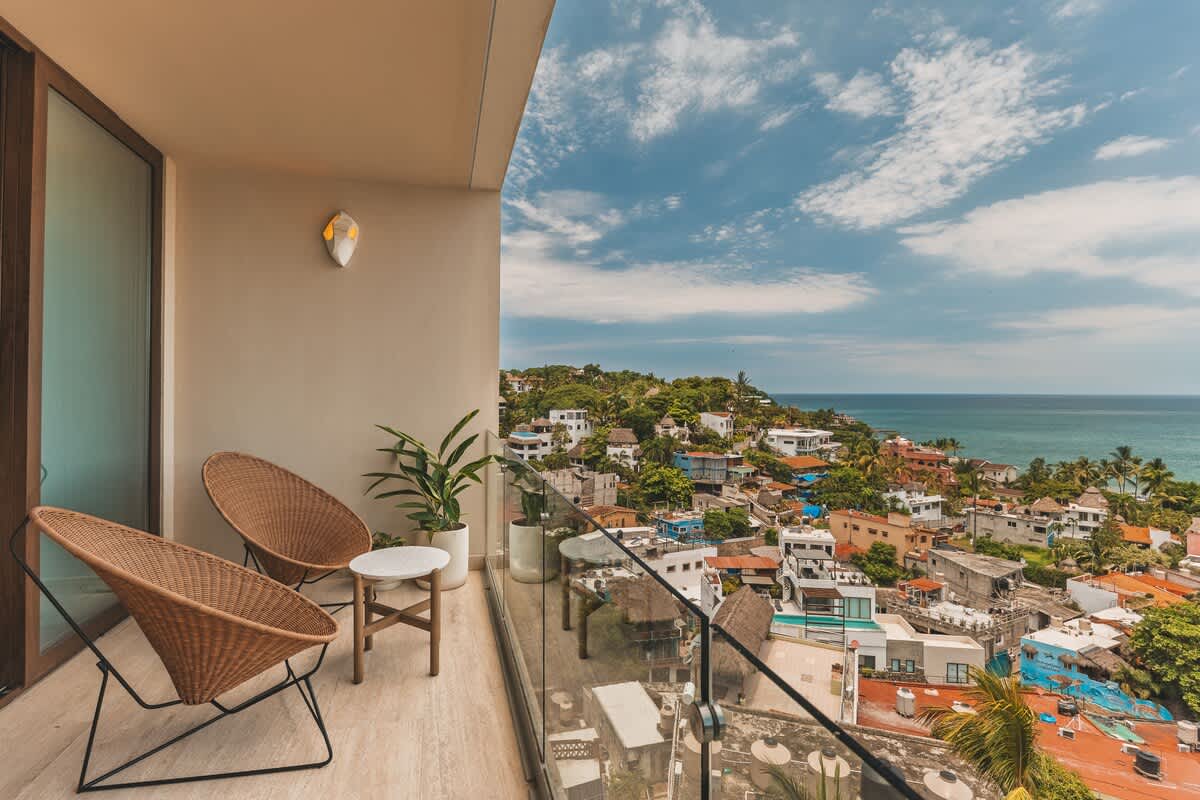 Wide shot of a balcony with two woven chairs and a small table, offering a sweeping view of a vibrant coastal town and the ocean, with a glass railing.