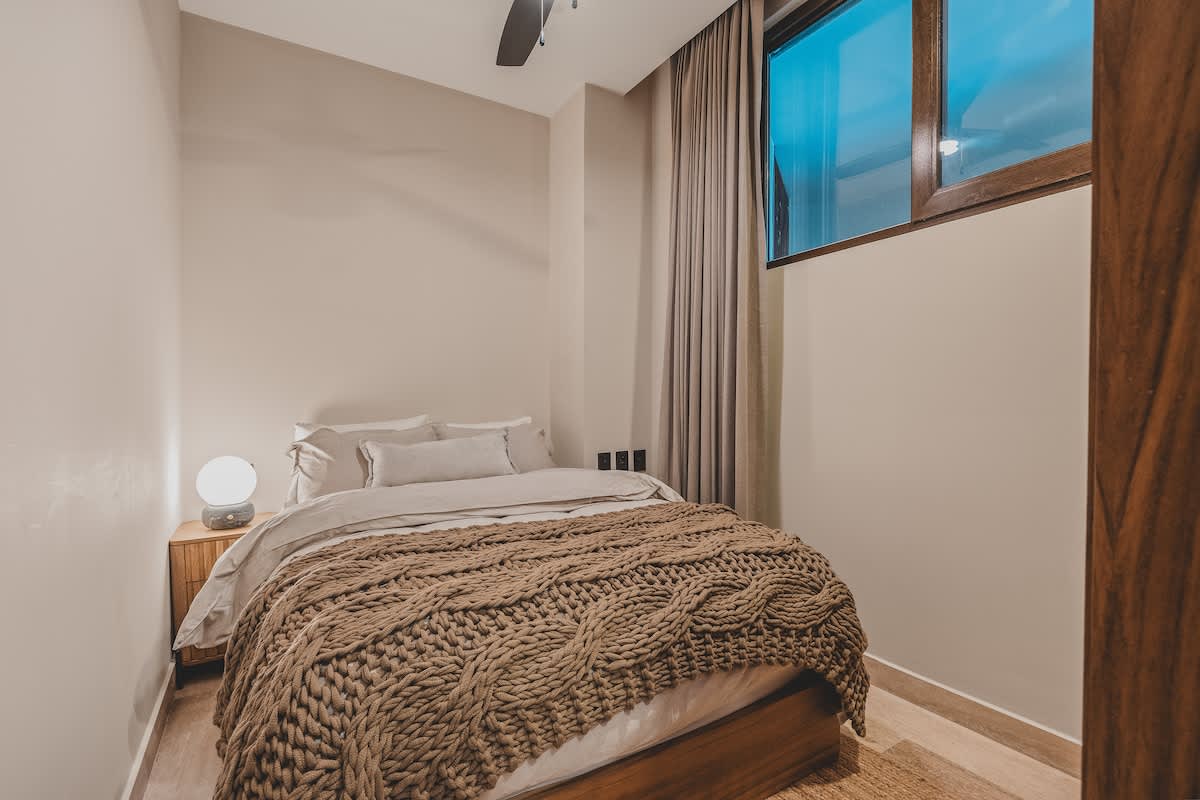 Another view of the cozy bedroom with a queen bed, a nightstand with a lamp, and a large window with curtains, showing a ceiling fan above.