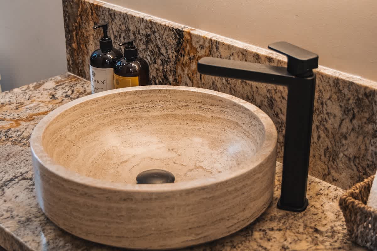 A modern bathroom vanity with a round, light-colored stone vessel sink and a matte black single-handle faucet, set on a speckled granite countertop with two dark pump bottles in the background.