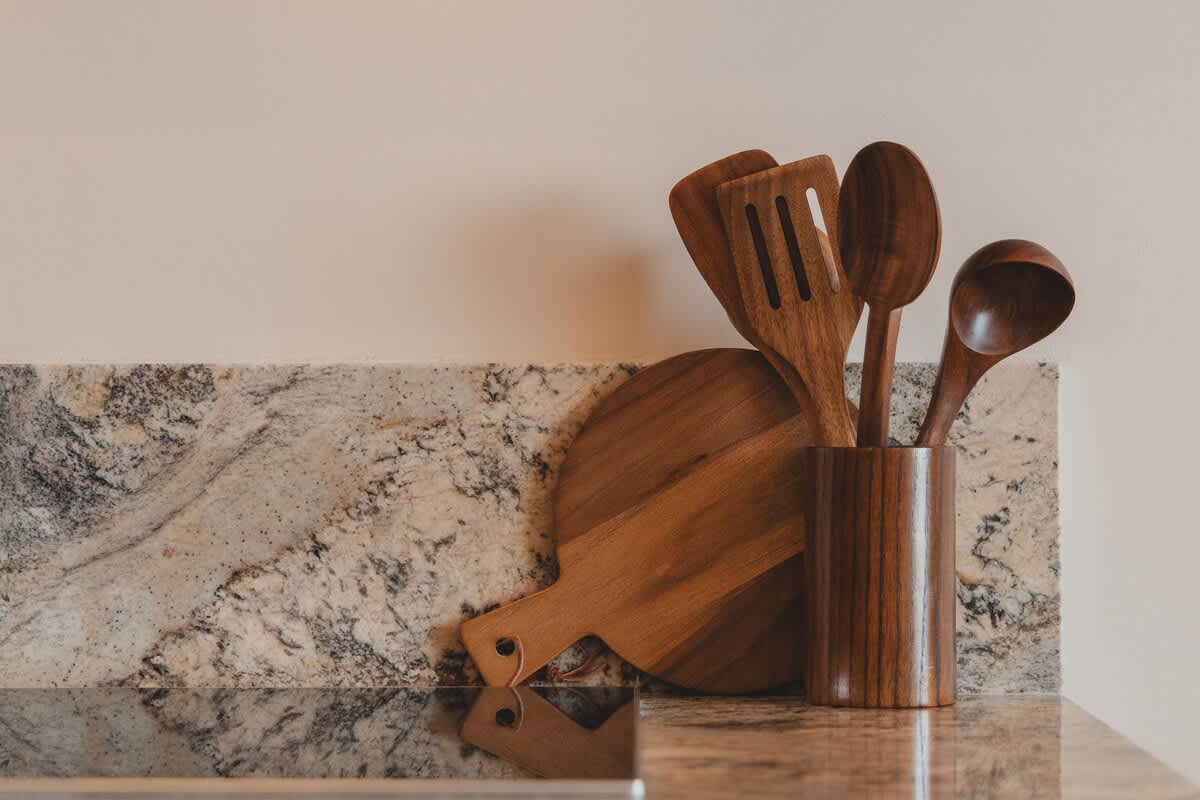 A close-up of a kitchen counter with a light brown granite surface and matching backsplash, featuring a wooden utensil holder filled with various wooden cooking tools. A round wooden cutting board leans against the wall, reflecting a warm, natural aesthetic.
