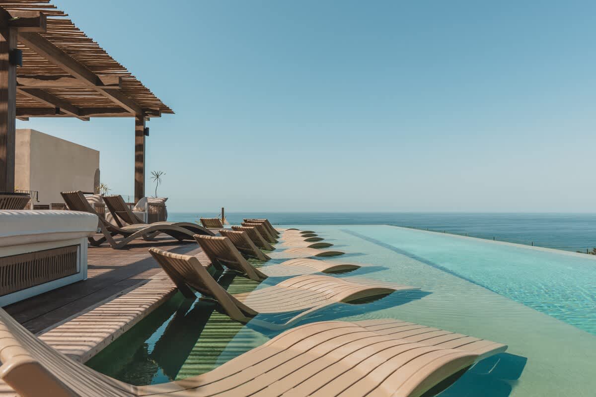 Panoramic view of the infinity pool with a line of submerged lounge chairs, bordered by a wooden deck and a shaded pergola, all overlooking the vast blue ocean.