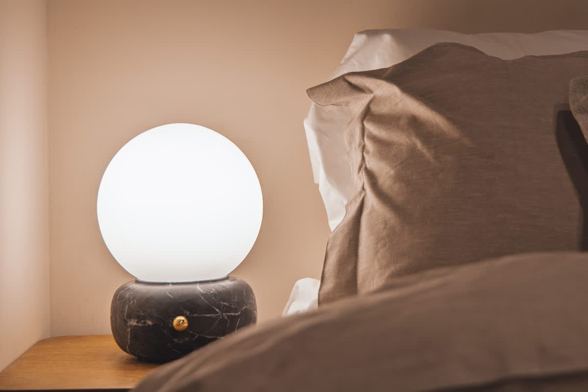 A cozy bedside scene featuring a modern spherical white lamp with a dark marble base on a wooden nightstand, next to a bed with light-colored pillows and bedding.