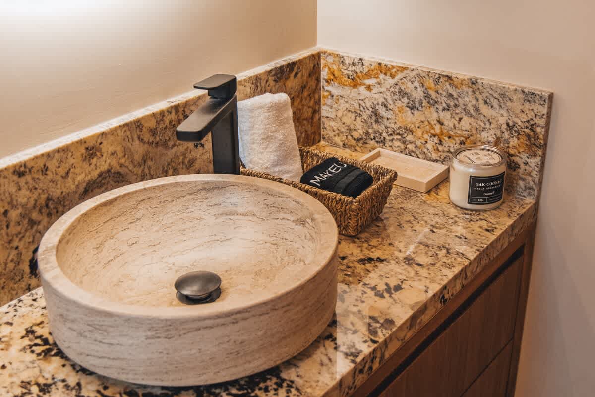 A stylish bathroom vanity with a round stone vessel sink, a matte black faucet, a woven basket with towels, and a white candle, all arranged on a speckled granite countertop with a wooden cabinet below.