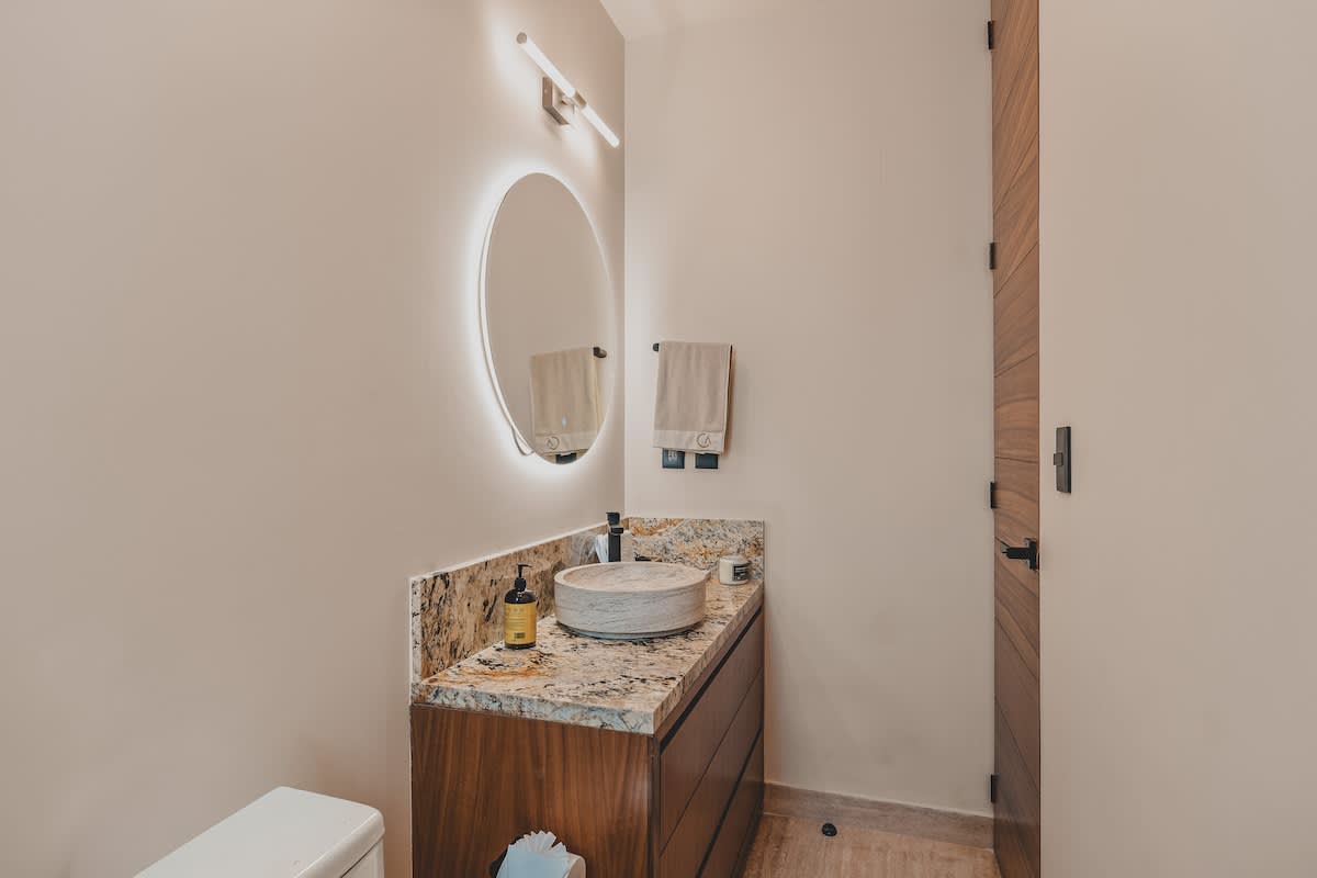 The bathroom vanity area showcases a round backlit mirror above a vessel sink set into a granite countertop. A dark wood vanity cabinet provides storage, with a toilet partially visible to the left.