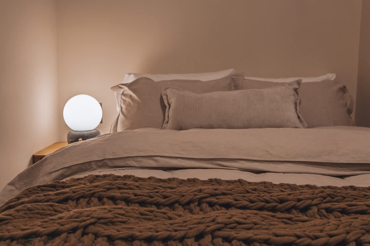 A comfortable bedroom featuring a bed with neutral-colored linens and pillows, accented by a chunky brown knitted throw blanket, with a spherical bedside lamp on a wooden nightstand.