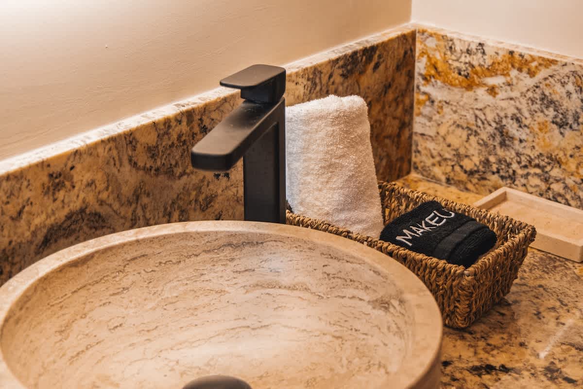A close-up of a contemporary bathroom vanity featuring a round stone vessel sink, a matte black faucet, and a woven basket with white and black "MAKEUP" towels on a speckled granite countertop.