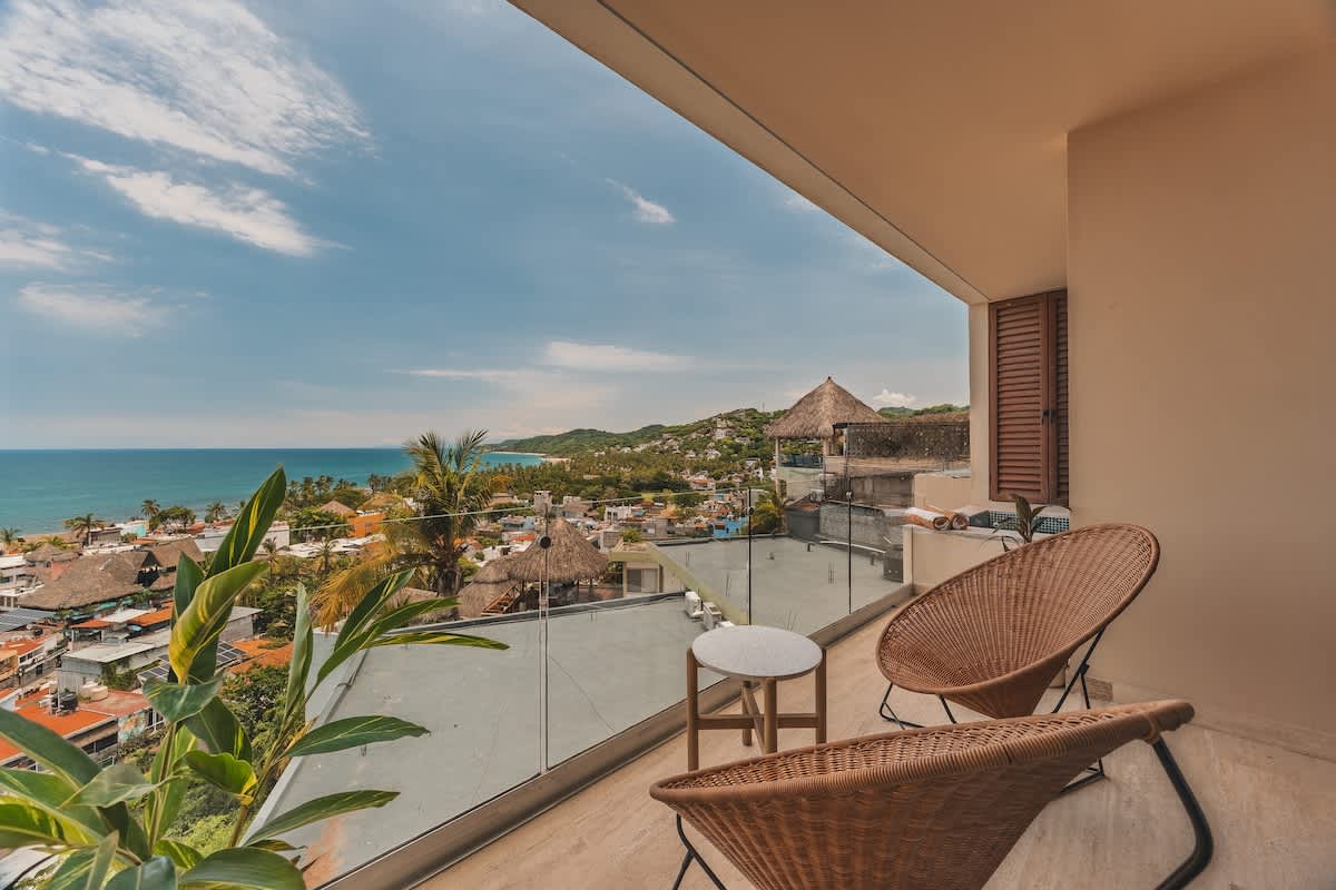 Angled view of a balcony with two woven chairs and a small side table, overlooking a picturesque coastal town and the ocean, with a glass railing providing an unobstructed vista.