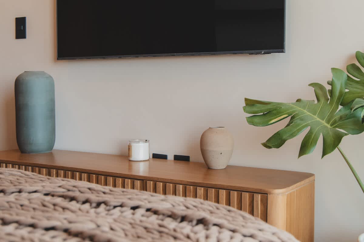 A modern bedroom setup with a wall-mounted TV above a sleek wooden console, adorned with two decorative vases and a candle, with a cozy knitted blanket in the foreground.