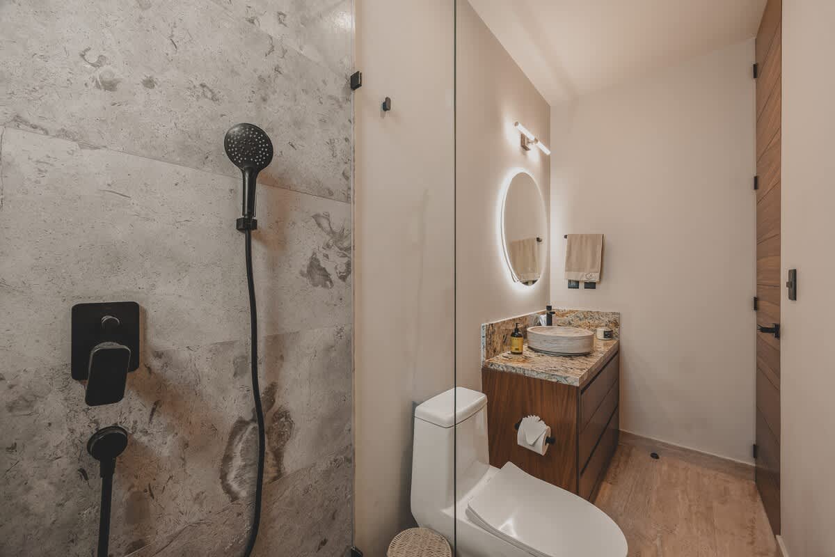 A modern walk-in shower with a black rain showerhead and handheld sprayer is adjacent to the toilet. The vanity with a vessel sink and backlit mirror is visible in the background, maintaining a cohesive design.