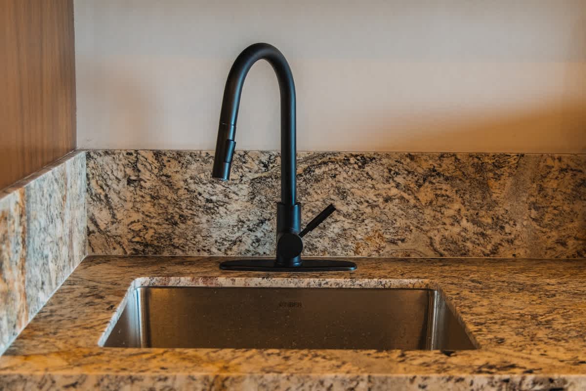 A stainless steel undermount sink with a modern matte black gooseneck faucet is set into a light brown granite countertop. The granite backsplash matches the counter, and a light-colored wall is visible above.