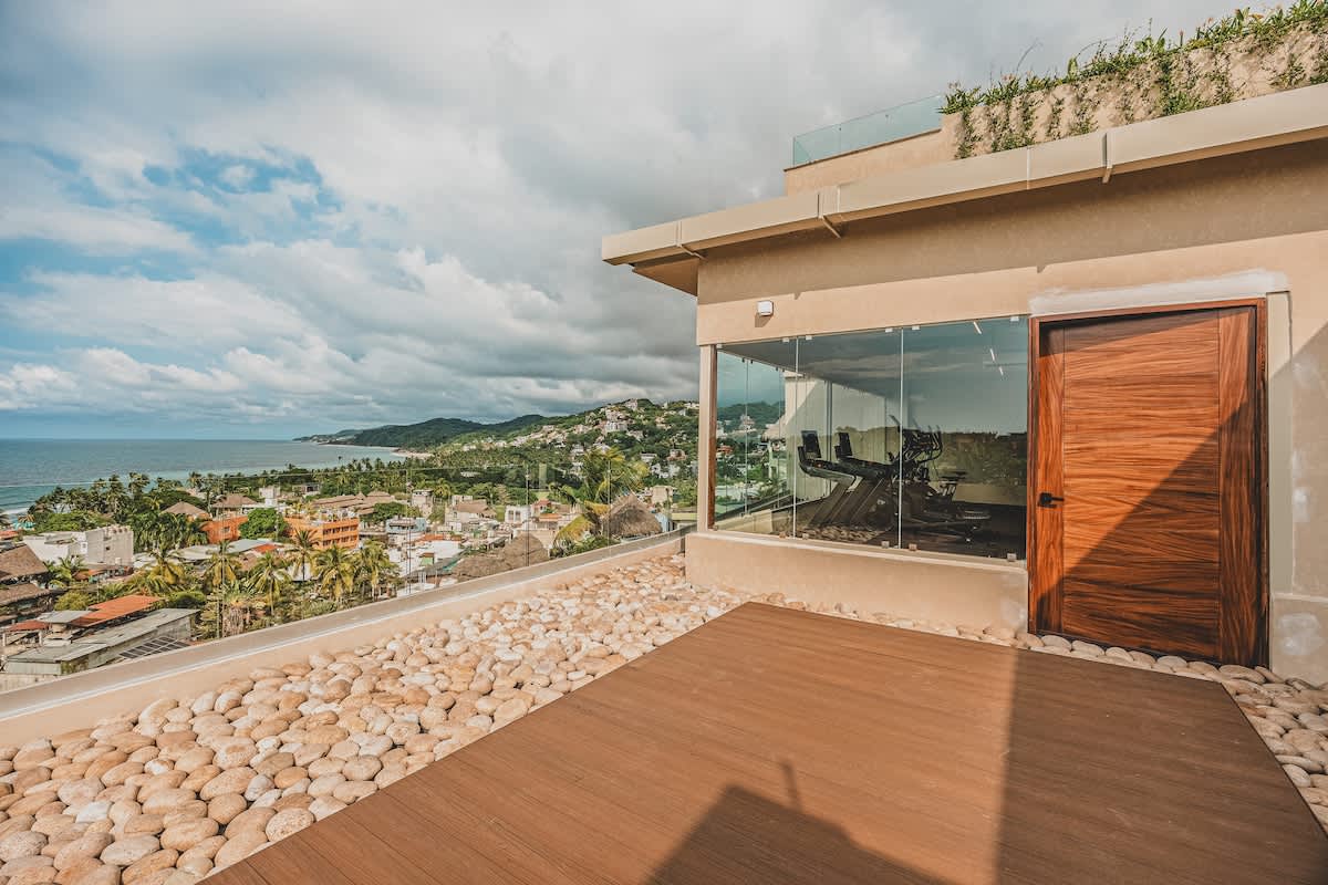 A spacious outdoor deck with wooden flooring and decorative river stones provides an expansive view of the ocean and a bustling coastal town, adjacent to a building with large glass windows revealing the interior gym. The sky is partly cloudy.