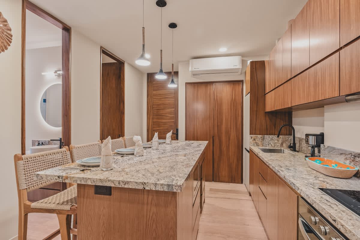 Stylish kitchen island serving as a dining area with bar stools, adjacent to a well-equipped kitchen with granite counters and wood cabinets, with a glimpse of a modern bathroom entrance.